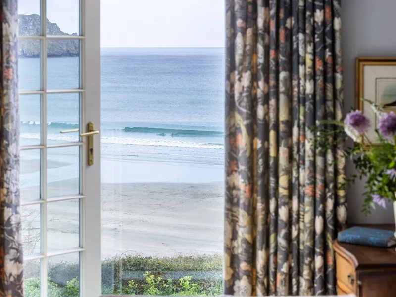 View through French doors towards a quiet sandy beach and calm sea, framed by dark floral curtains with flowers on a wooden table inside.