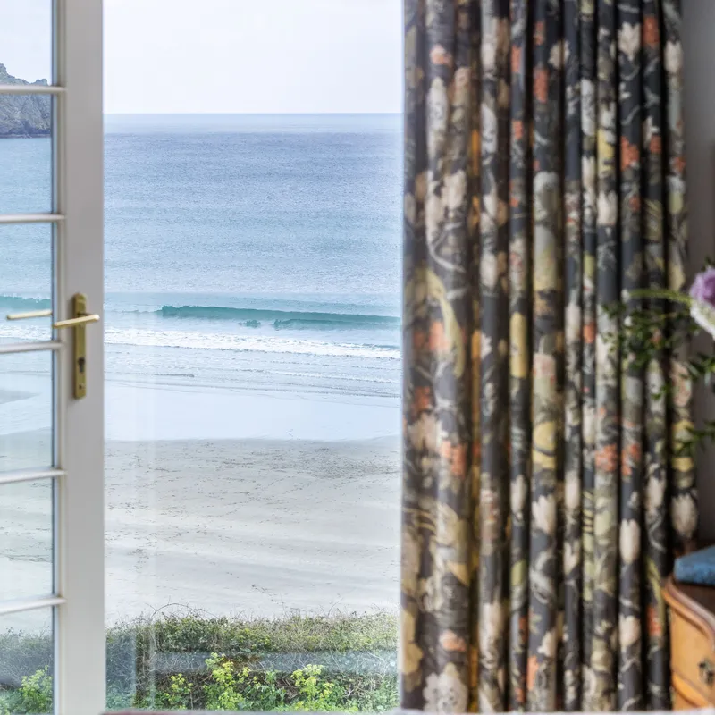View through French doors towards a quiet sandy beach and calm sea, framed by dark floral curtains with flowers on a wooden table inside.