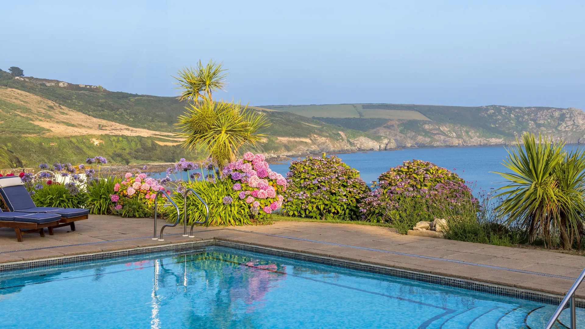 Outdoor swimming pool with sun loungers, colourful flowering garden and sweeping coastal sea views