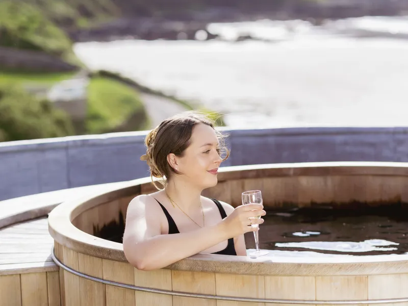 Woman relaxing in a wooden hot tub with a glass of champagne overlooking a coastal bay