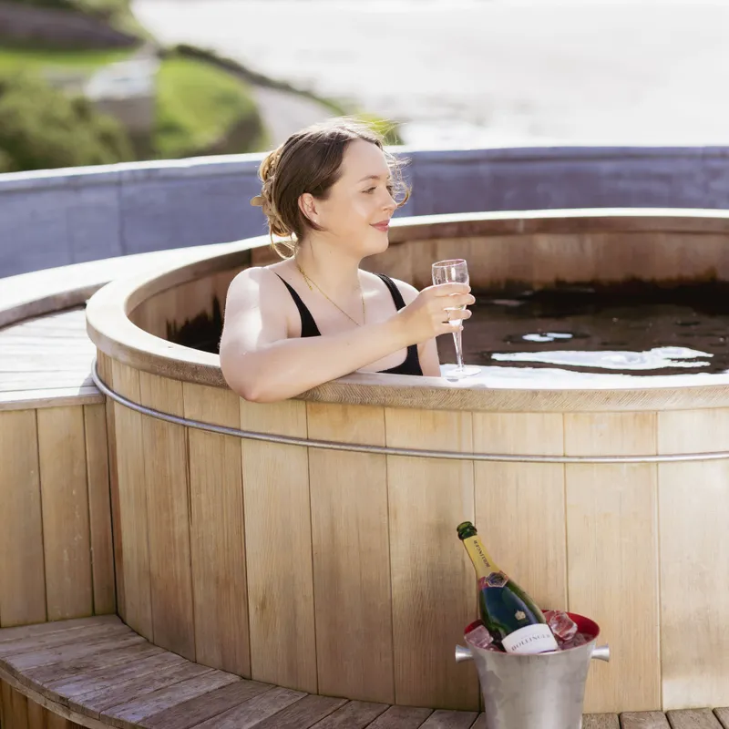 Woman relaxing in a wooden hot tub with a glass of champagne overlooking a coastal bay