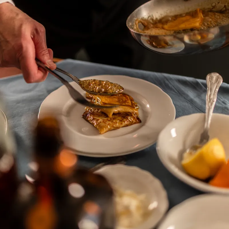Chef spooning warm sauce over plated crêpes at a dining table