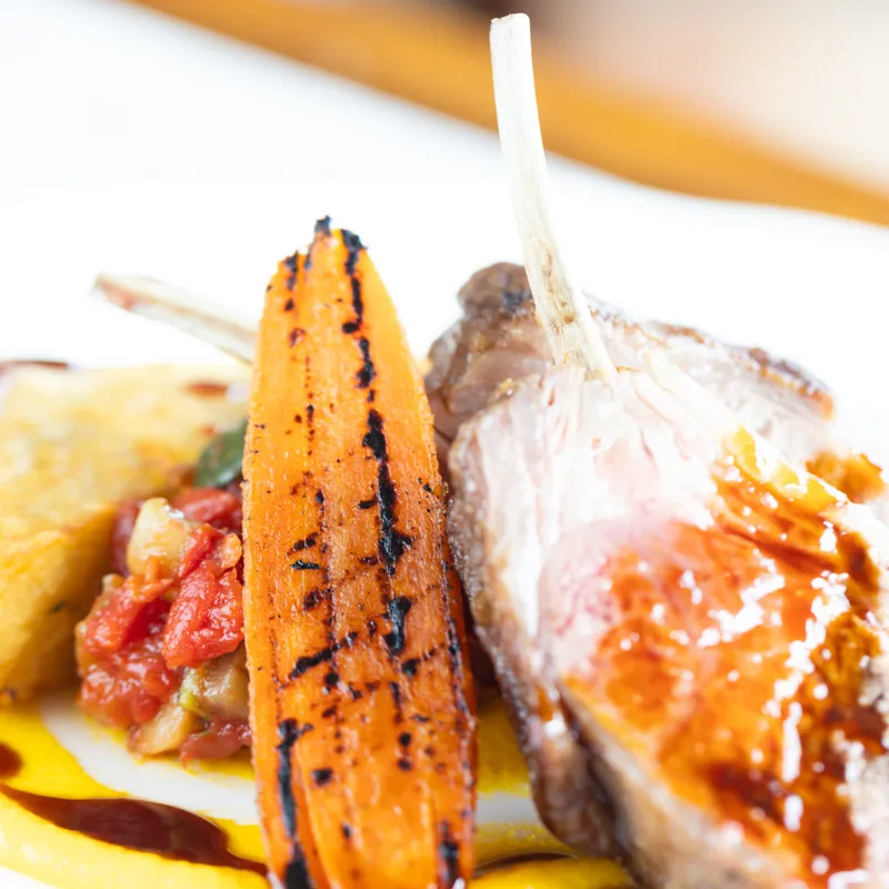 Close-up of lamb cutlets with a glazed finish, served with charred baby carrots, creamy yellow purée and a vegetable garnish.