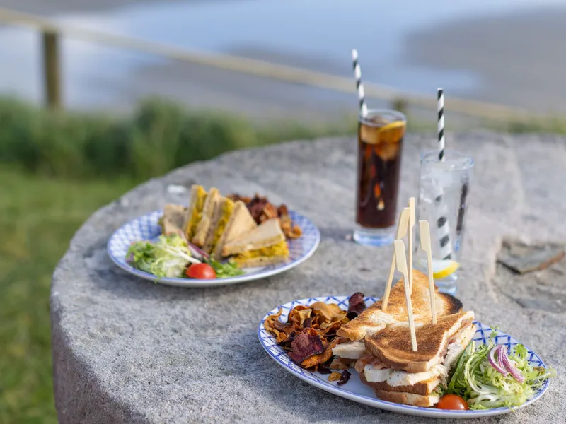 Plates of toasted sandwiches and drinks on a stone table overlooking a sandy beach and blue sea.