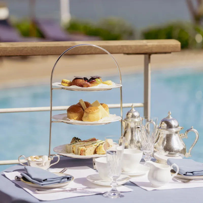 Afternoon tea with sandwiches, scones and pastries served on a tiered stand beside a pool with coastal views in the background