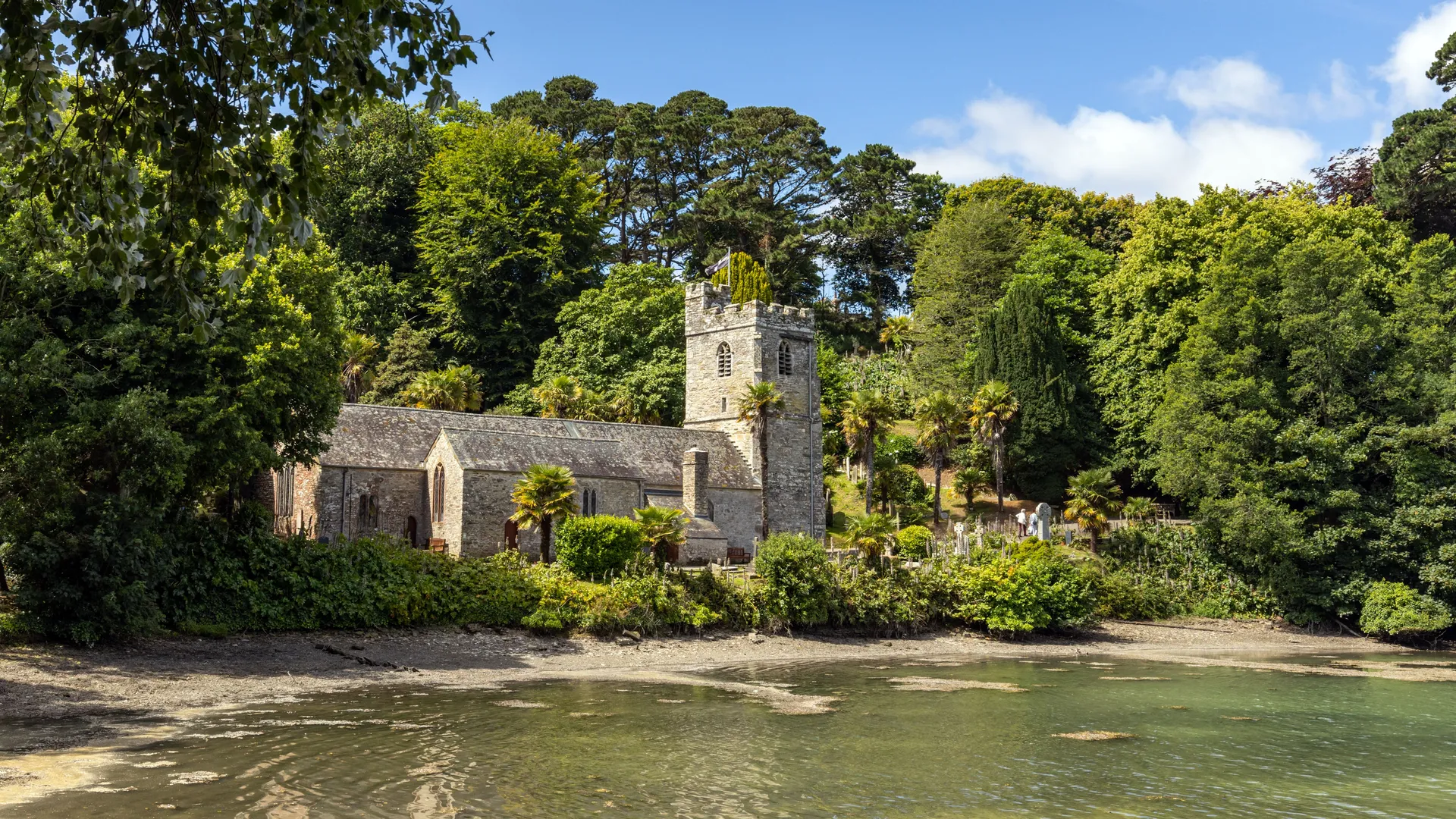 St Just in Roseland Church with a square tower beside a calm tidal creek, surrounded by dense green trees under a bright blue sky.