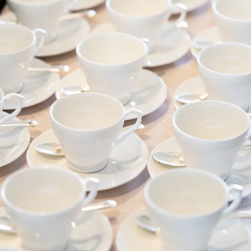 Rows of white teacups and saucers with teaspoons arranged on a table