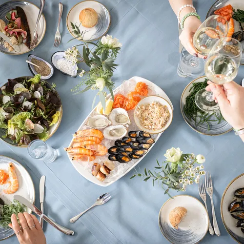 Overhead view of a seafood dining table with oysters, prawns, mussels, salad and glasses of white wine being raised for a toast