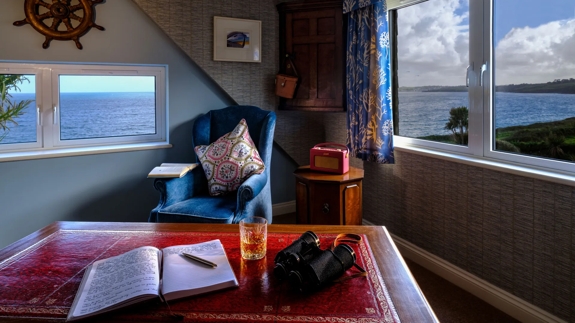Cosy seaside study with a blue armchair, writing desk, binoculars and sea views through two windows.
