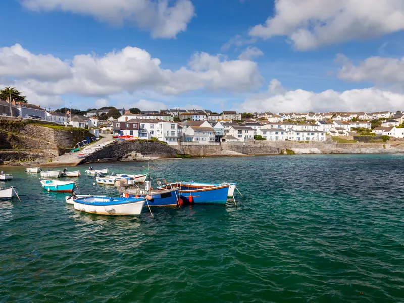 Portscatho Cornwall Small fishing boats moored in a sheltered harbour with seaside houses and stone walls under a bright blue sky with scattered clouds.