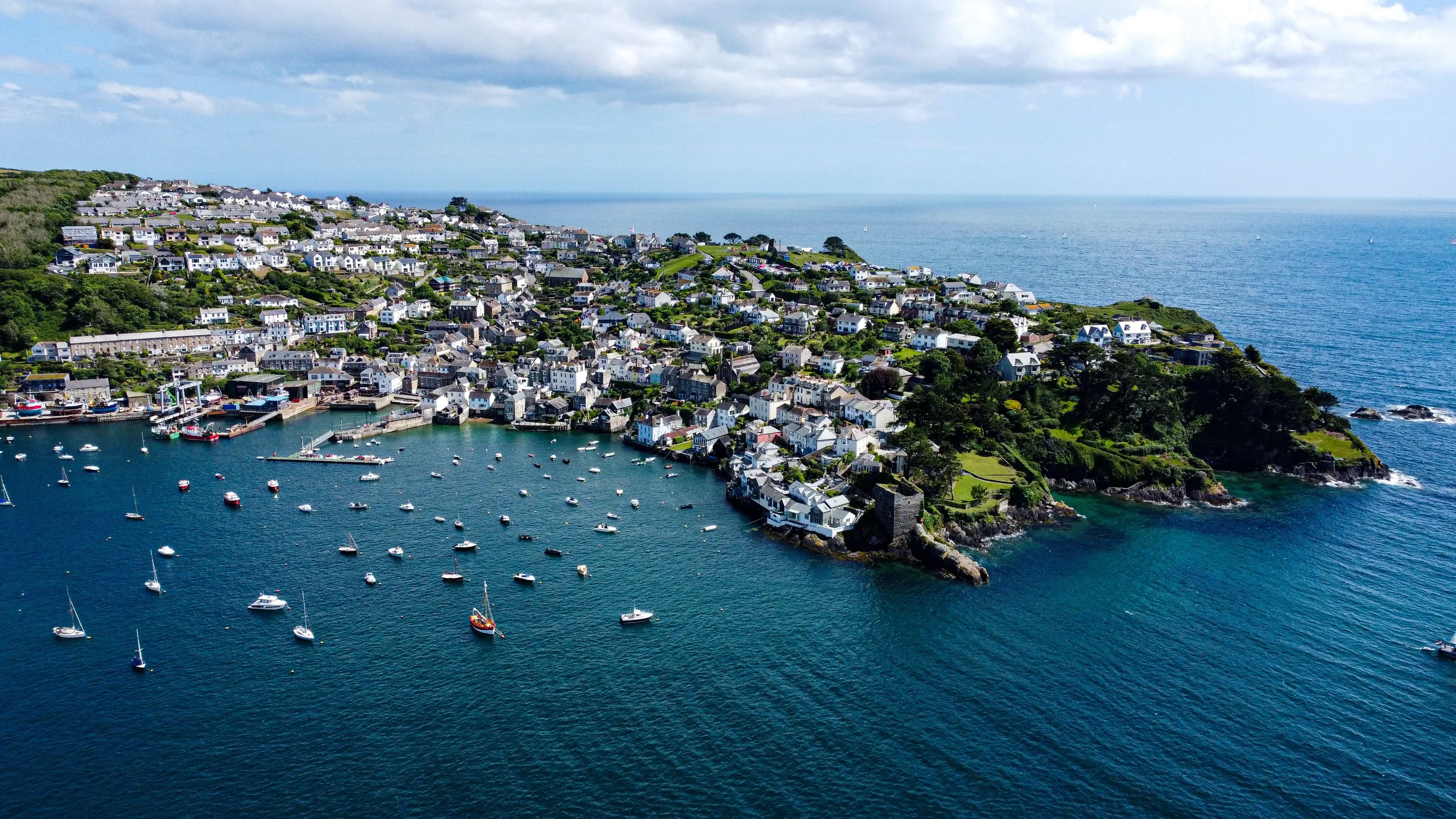Aerial view of a coastal village with a small harbour, moored boats and houses clustered on a green headland beside the sea.