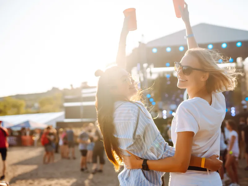 Two young women wearing sunglasses embrace and raise drinks at an outdoor music festival in bright sunshine.
