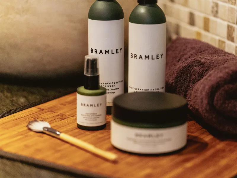 Bramley toiletries arranged on a wooden tray beside a rolled towel in a bathroom