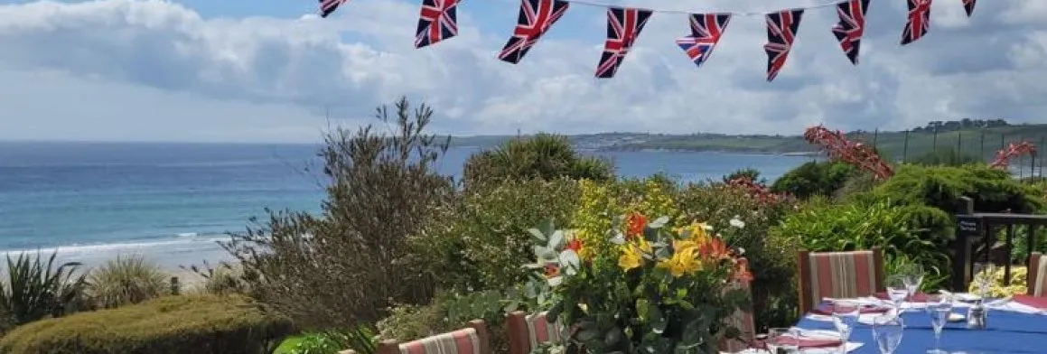 Outdoor dining table set for a celebration with Union Jack bunting, overlooking a coastal garden and sea view.