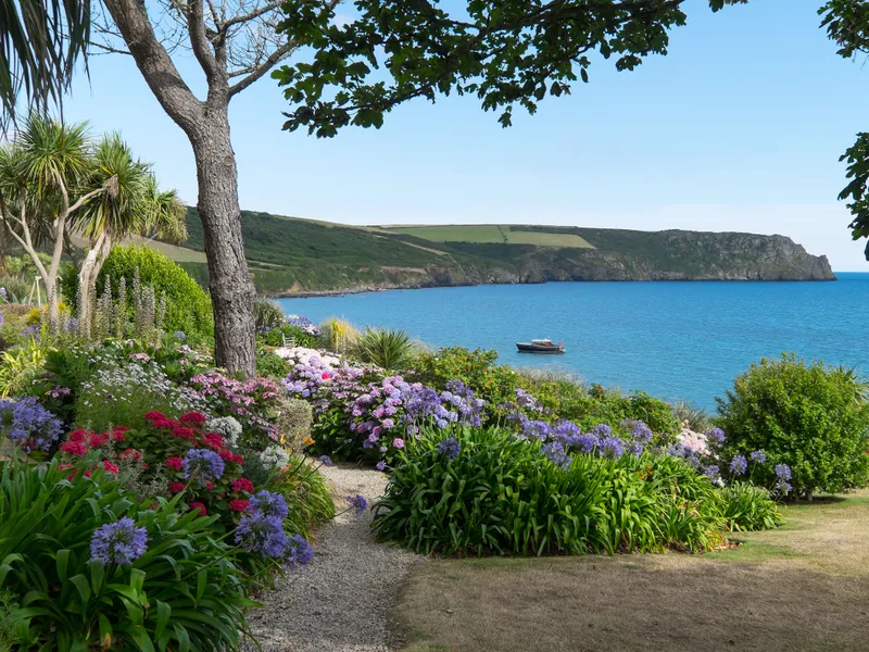 The Nare's flower-filled coastal garden overlooking a calm blue bay with a small boat and rugged headland beyond