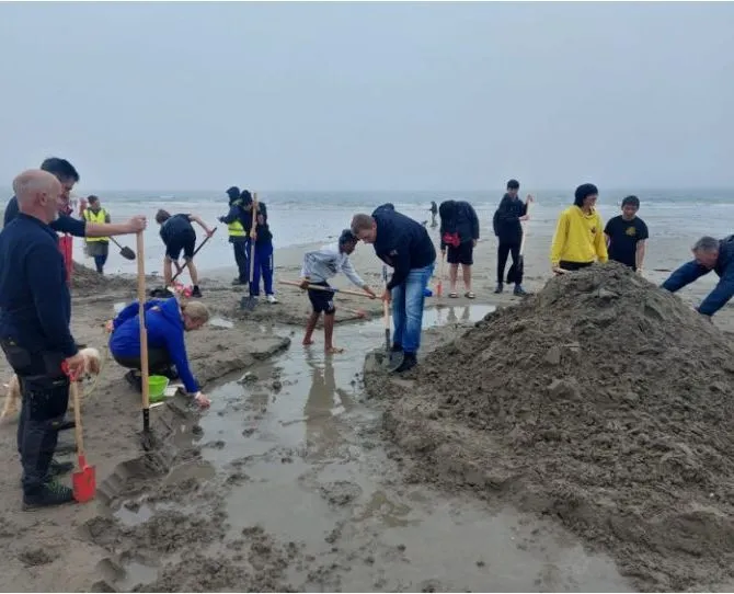 Group of people digging channels and moving sand on a beach with spades on an overcast day