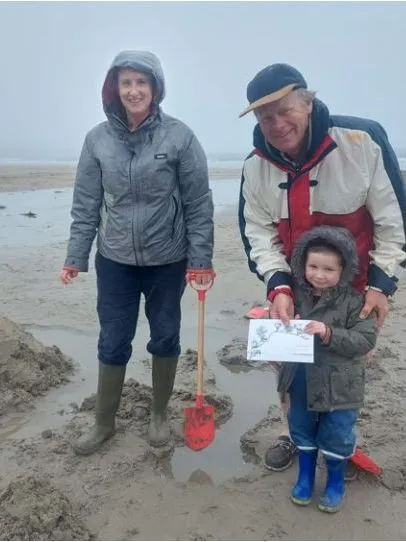 Family on a misty beach beside a sandcastle with a Union Jack flag, with a child holding a certificate