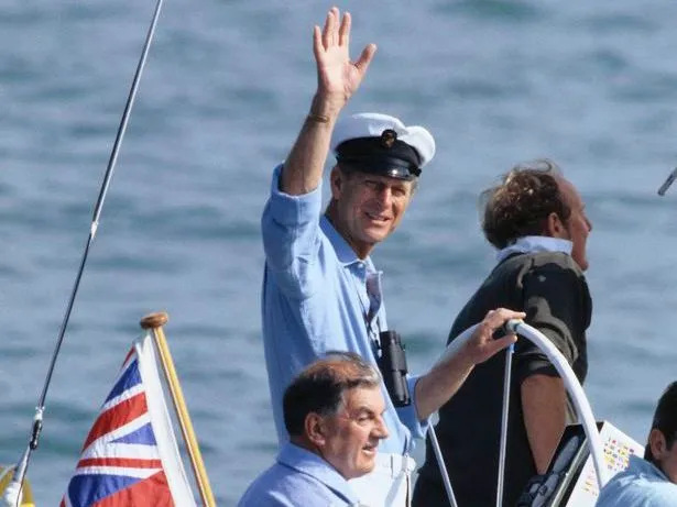 A man in a sailor’s cap waves from a boat, with a Union Jack flag visible and the sea in the background.