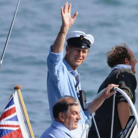 A man in a sailor’s cap waves from a boat, with a Union Jack flag visible and the sea in the background.