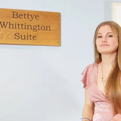 Woman standing beside a wooden plaque reading 'Bettye Whittington Suite' in an interior room.