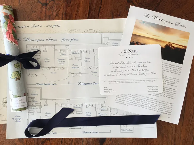 Printed launch materials for The Whittington Suites at The Nare, including site and floor plans, a brochure, an invitation card and a floral rolled document tied with a navy ribbon on a woode