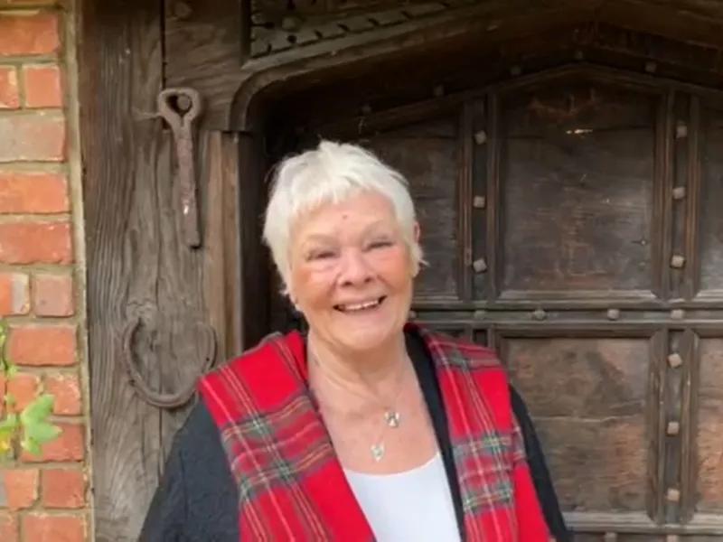 Dame Judi Dench smiling in front of a rustic wooden doorway and brick wall, wearing a red tartan shawl.