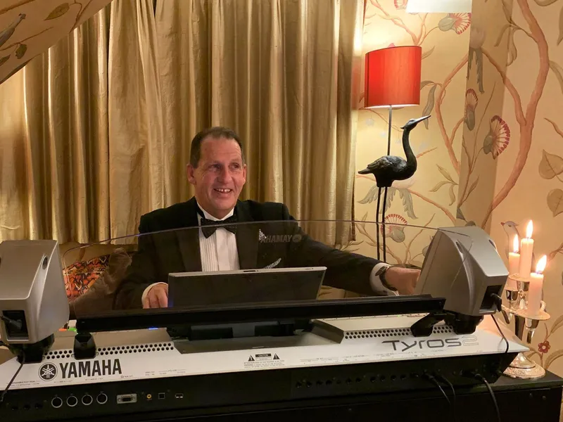 A man in a black tie dinner jacket playing a Yamaha keyboard in an elegant room with floral wallpaper, red lampshades and candlelight.