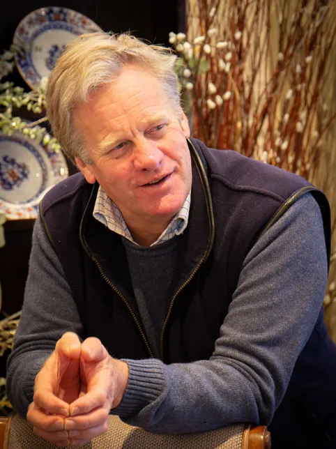 Portrait of a smiling man seated indoors, leaning forward with clasped hands against a warm decorative background.