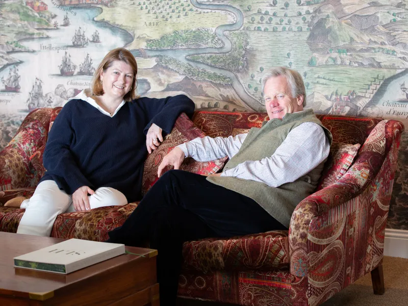 Two staff members seated on a patterned sofa in front of a large antique-style map mural, smiling at the camera.