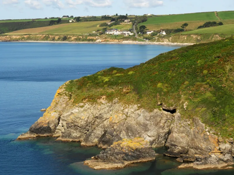 Rocky coastal headland with green cliffs overlooking calm blue sea and patchwork fields in the distance