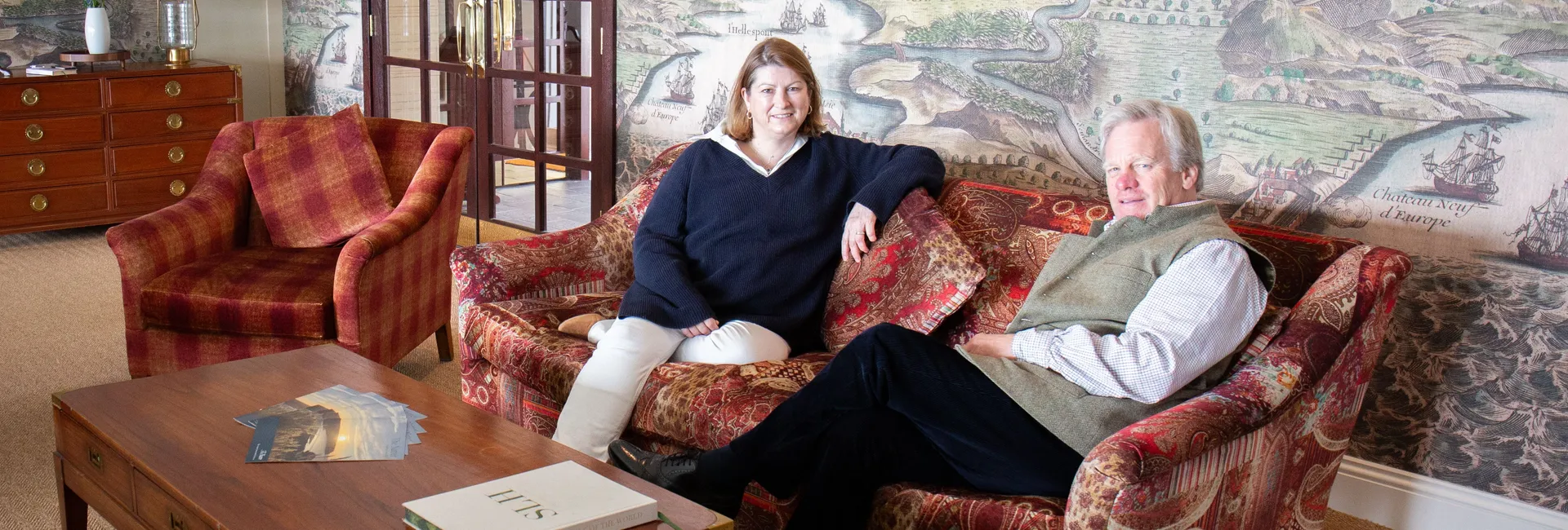 Two staff members seated on a patterned sofa in an elegant lounge with a large mural map on the wall behind them.