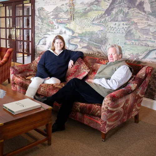 Two staff members seated on a patterned sofa in an elegant lounge with a large mural map on the wall behind them.