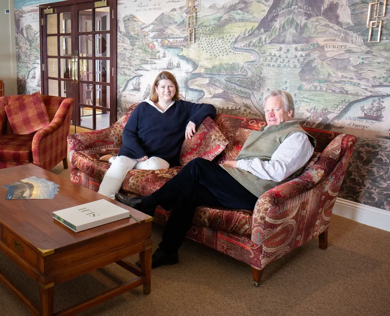 Two staff members seated on a patterned sofa in an elegant lounge with a large mural map on the wall behind them.