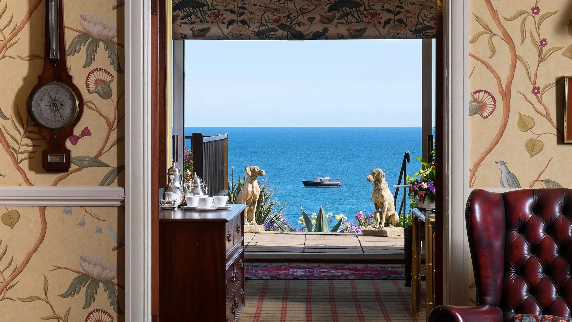 Elegant reception room with floral wallpaper, antique furnishings and an open doorway framing a sea view with a boat offshore