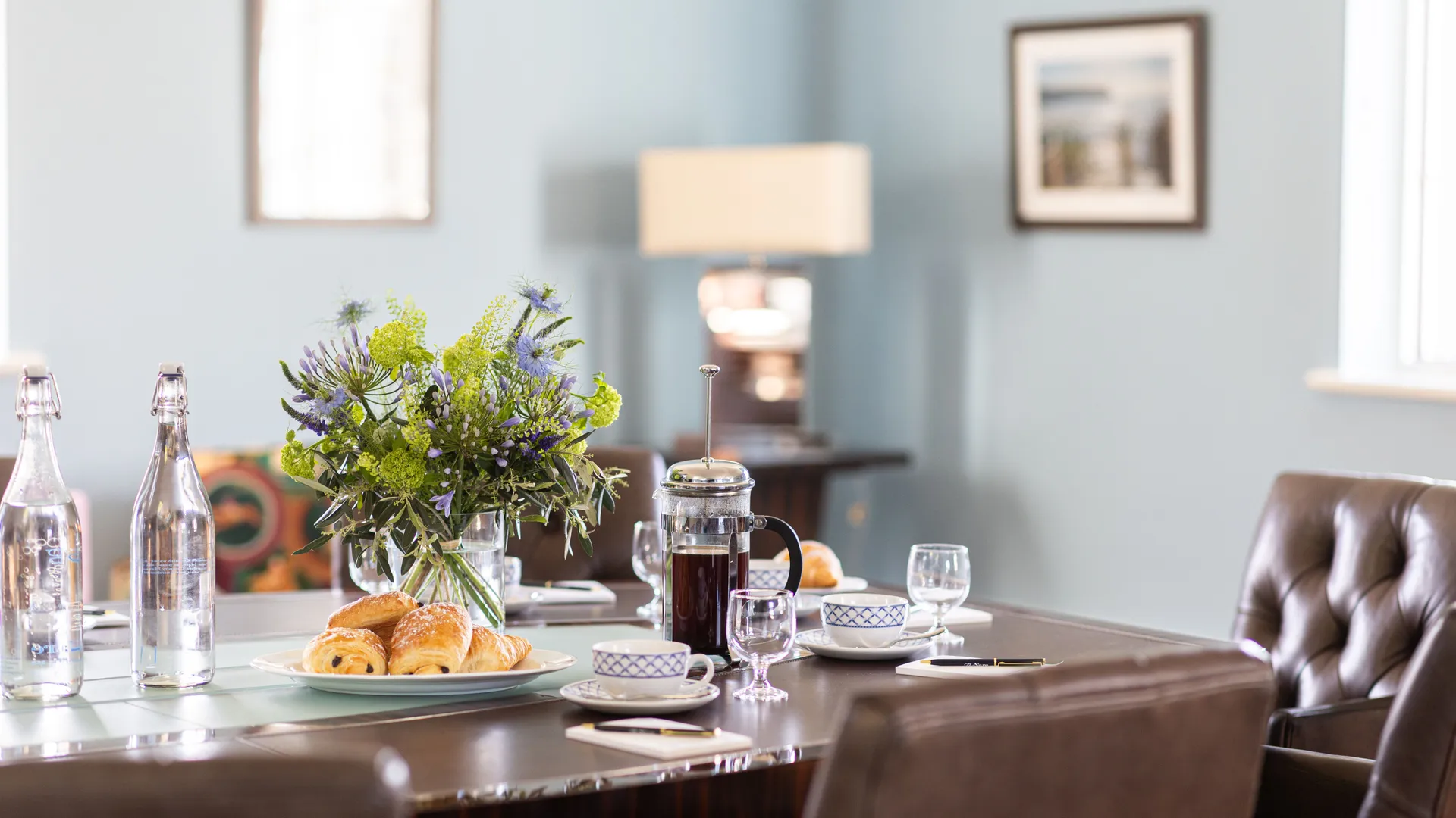 Meeting table set with coffee, pastries, water bottles and a fresh flower arrangement in a bright room