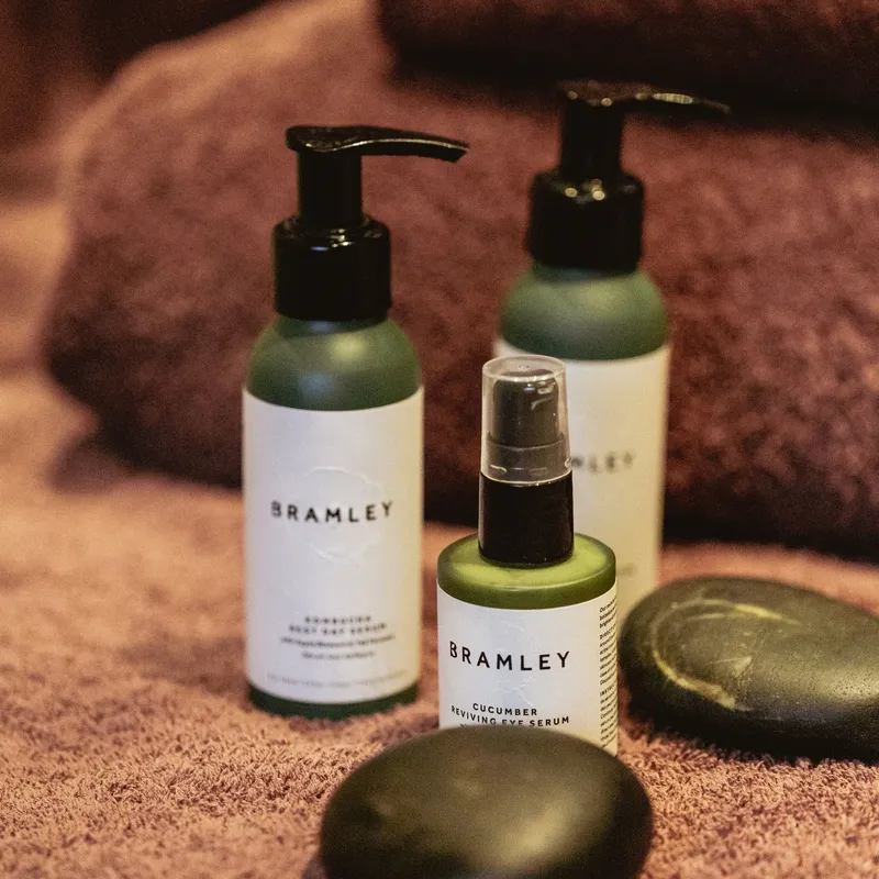 Bramley skincare bottles and cucumber reviving eye serum arranged on mauve towels with smooth massage stones