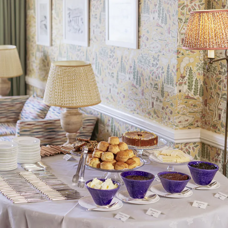 Afternoon tea table with scones, cakes, cream and jam in a cosy room with patterned wallpaper and lamps.