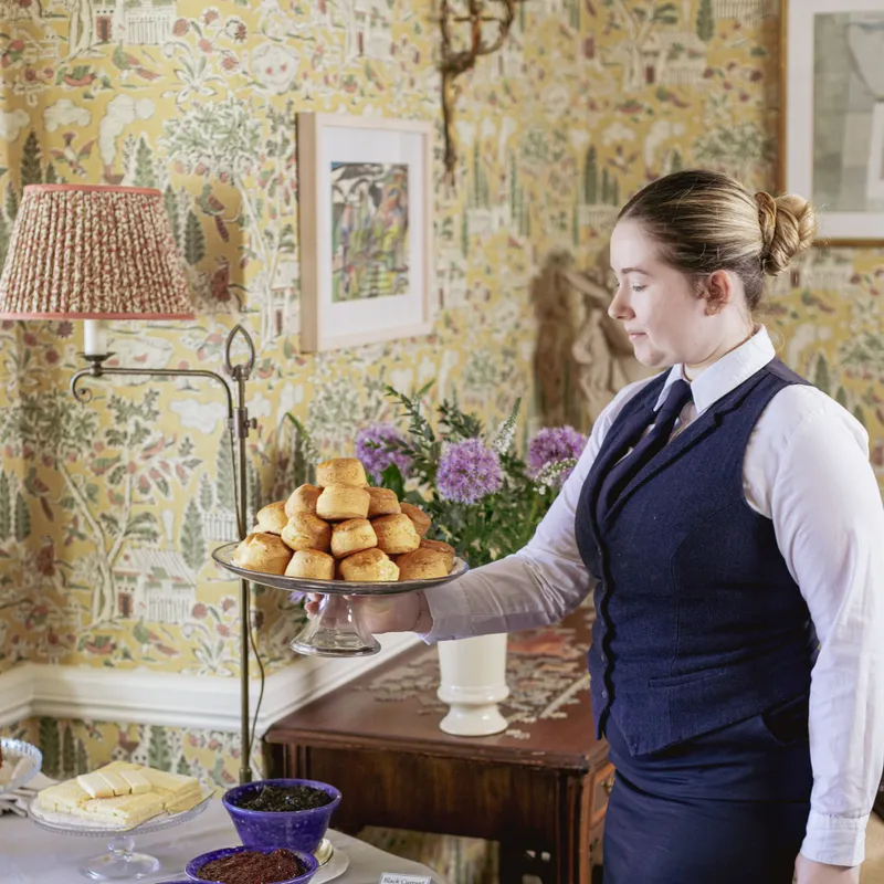 Staff member in formal uniform serving a tray of freshly baked scones beside a table with clotted cream and assorted jams