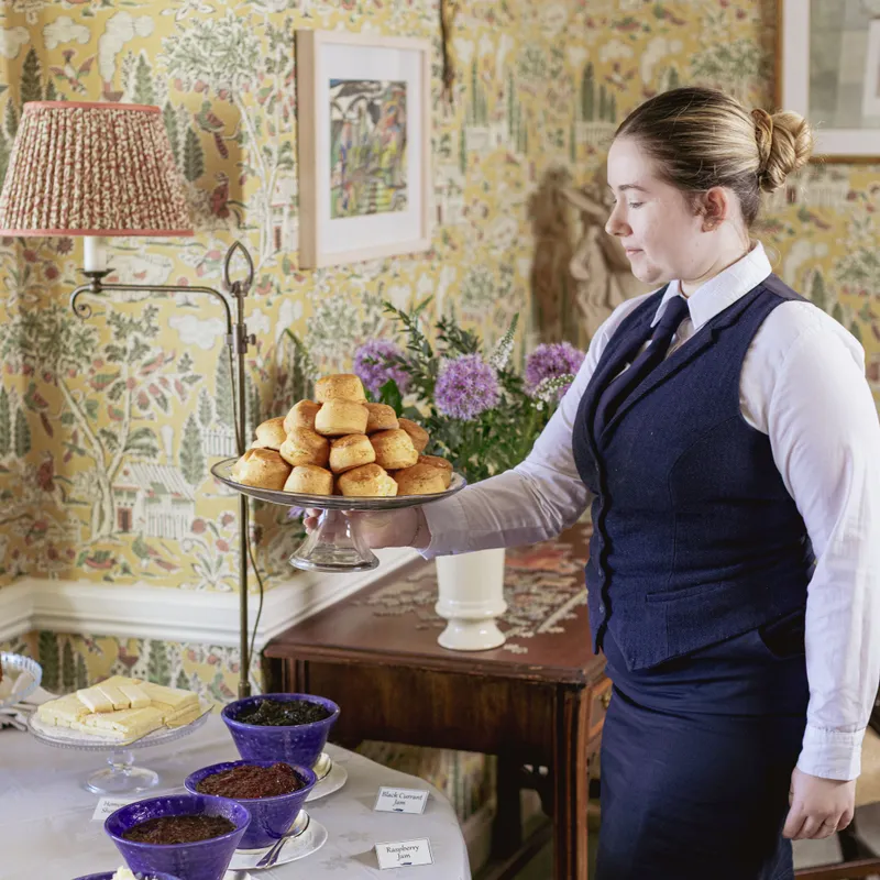 Staff member in formal uniform serving a tray of freshly baked scones beside a table with clotted cream and assorted jams