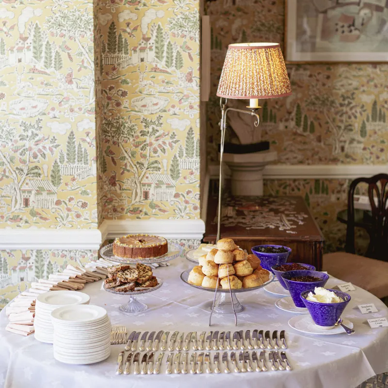 Afternoon tea table with scones, cakes and preserves set against patterned yellow wallpaper in a traditional interior