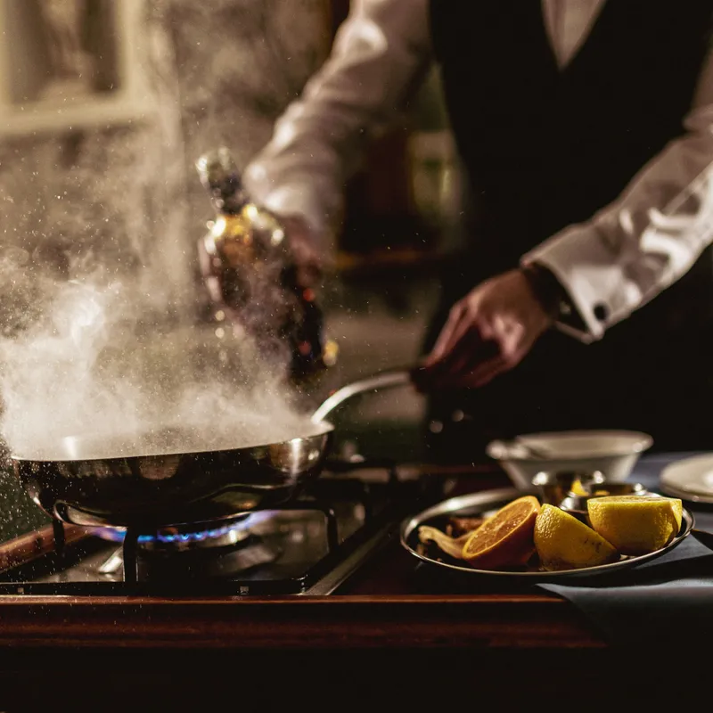 Chef preparing a flambéed dish in a steaming pan beside citrus wedges and a laid dining table