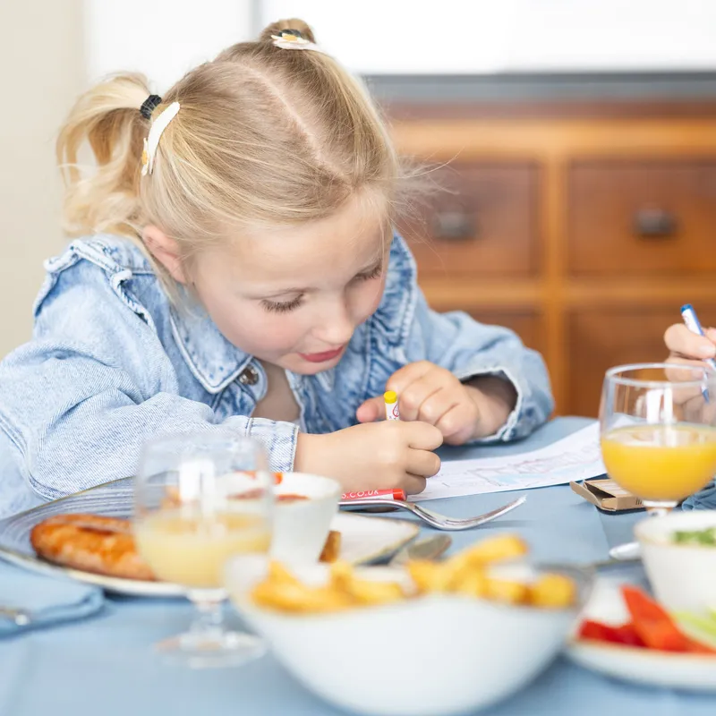 Young girl drawing with crayons at a dining table during a meal, with juice, chips and vegetables in front of her.