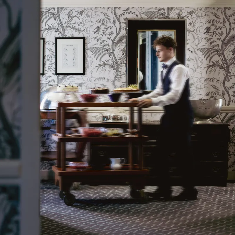 Blurred waiter pushing a dessert trolley through an elegant dining room with botanical wallpaper