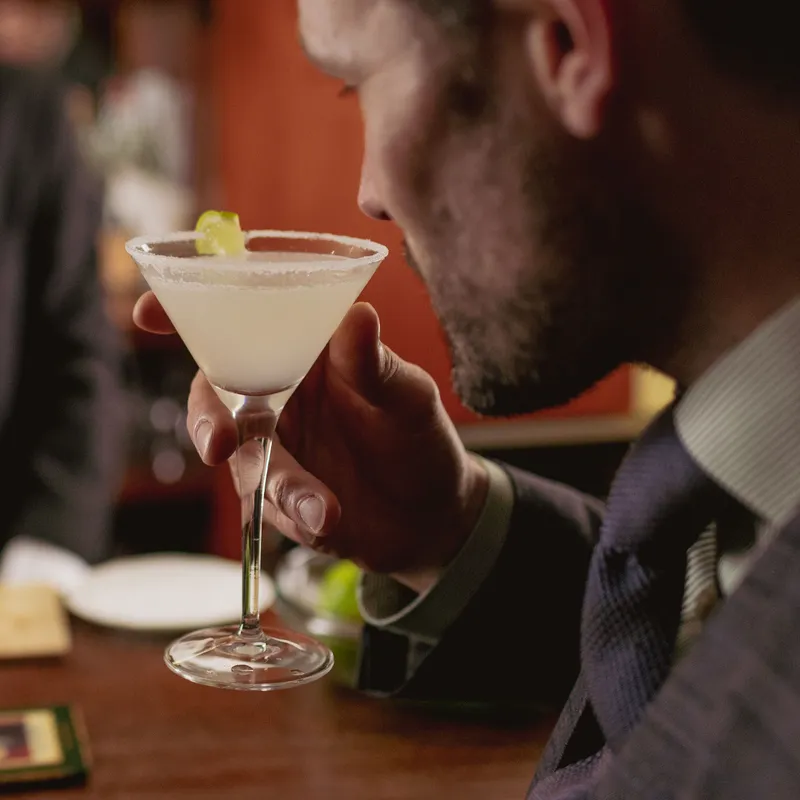 Man in a suit holding a pale cocktail in a martini glass at a bar