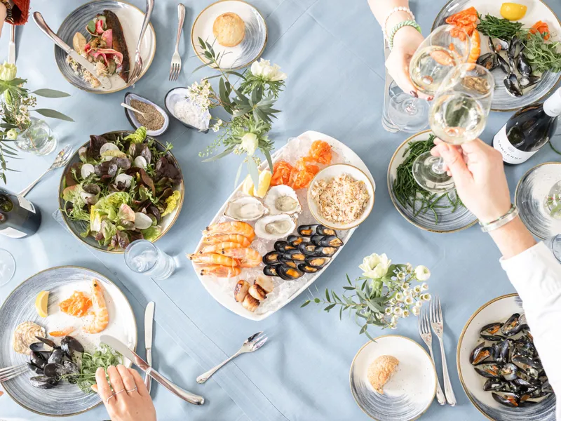 Overhead view of a seafood meal with prawns, oysters, mussels and salad on a pale blue table as guests raise glasses of white wine.