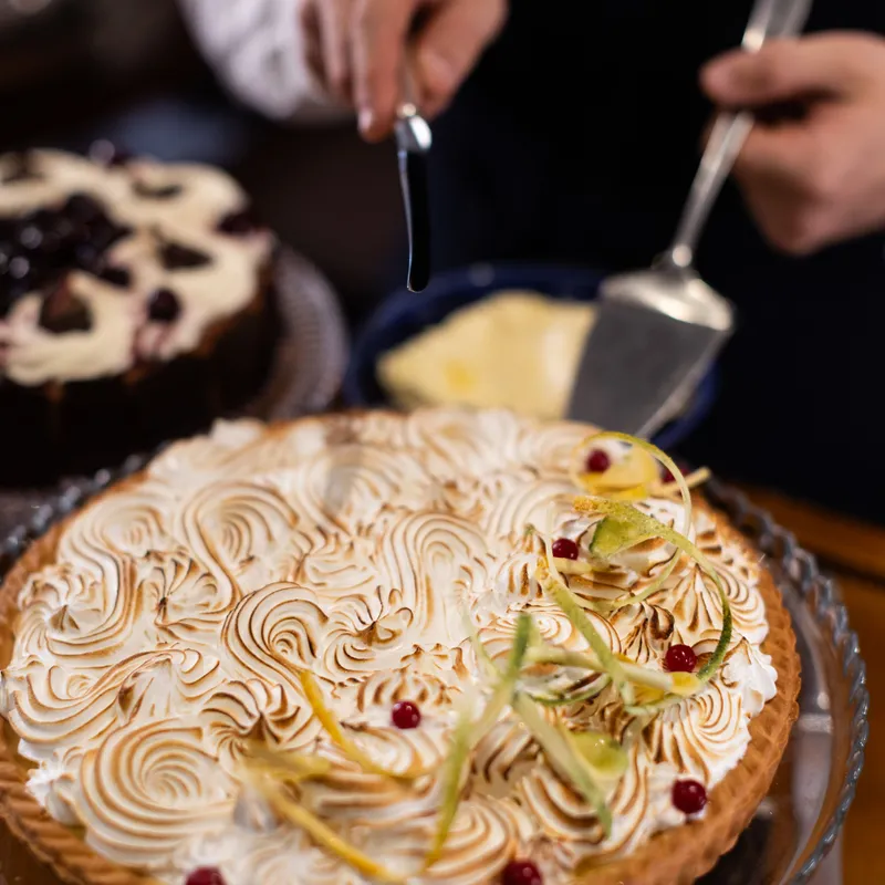 A toasted meringue tart garnished with citrus peel and red berries, with hands poised to serve it at a table.