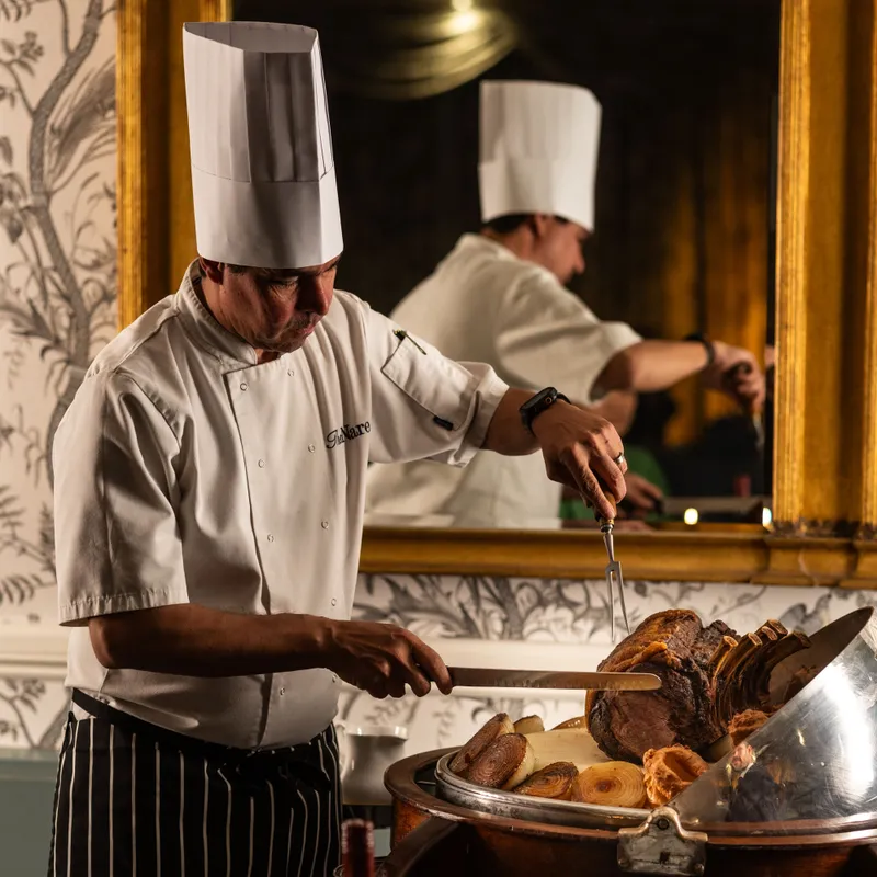 Chef carving a roast joint on a silver trolley in an elegant dining room with a large gilt mirror behind him.