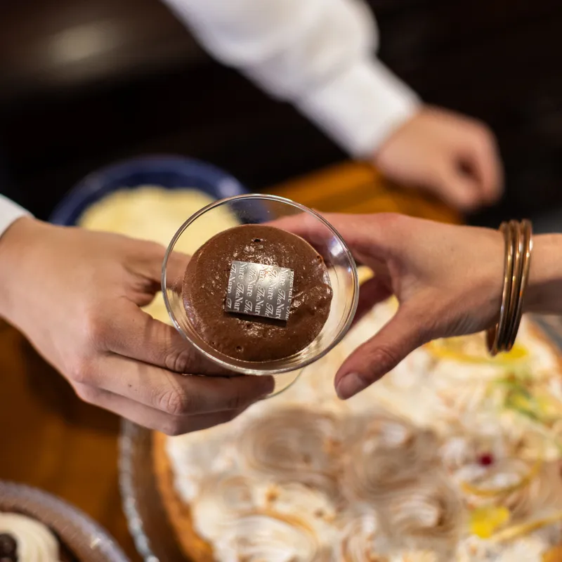 A person receives a glass dessert cup topped with chocolate over a table of assorted sweets.