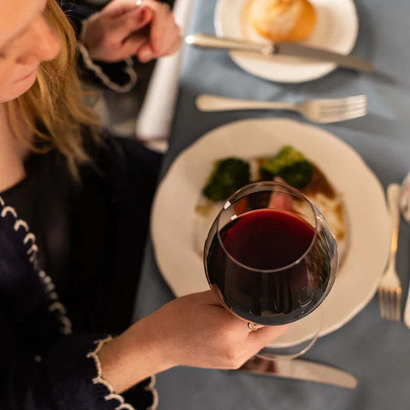 Woman holding a glass of red wine while dining at a table with a plated meal and bread roll.