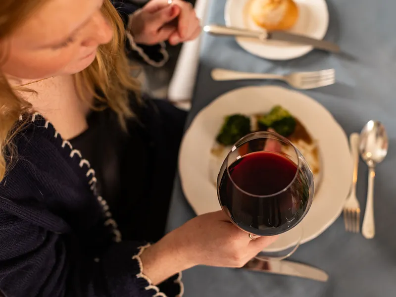 Woman holding a glass of red wine while dining at a table with a plated meal and bread roll.